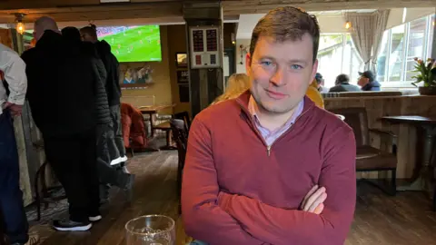 Man in pub in red sweater arms folded sits infront of pint on the table