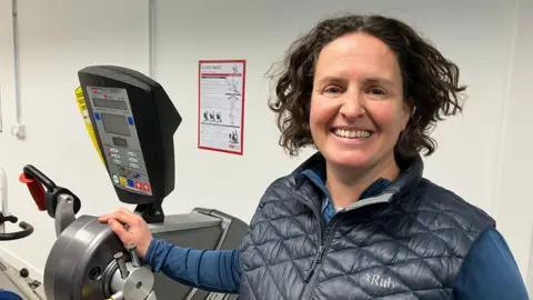 Kim has brown wiry hair and is smiling at the camera. She is wearing a 'Rab' navy gilet over a blue training top. Her hand is placed on one of the exercise machines. 