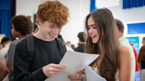 Getty Images A boy and girl open their exam results. They are smiling and holding white pieces of paper. They are in a hall and there are other students in the background. He is wearing a black jumper with a rucksack over his shoulders. The girl has long brown hair and is wearing a white vest.