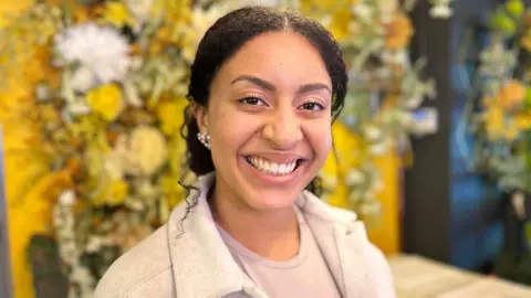 Khiefa Gabbidon A woman with tied-back black hair wearing an earring in her right ear, a light-purple shirt and a white jacket, is smiling while standing in front of a flower-covered wall in a restaurant.