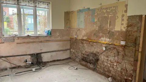 An image of the interior of a house with the walls stripped back to bare brick and a window in the left of the image. 