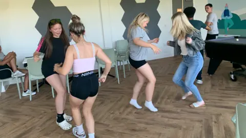 Debbie Etheridge Four young women in sports gear dancing in pairs in a community centre room with a parquet floor. There are other young people in the background, near a pool table