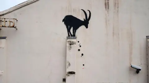 PA Media Banksy stencil piece showing goat perched on pillar on the side of a wall with rocks falling and a real CCTV pointed in its direction.