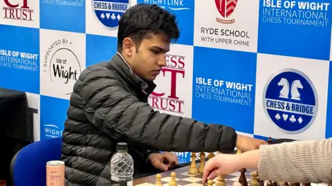 Shreyas, a teenage boy with thick dark hair, reaches over a chess board. He is wearing a black coat and sat in front of a sign showcasing various sponsors of the Isle of Wight Chess Tournament. 