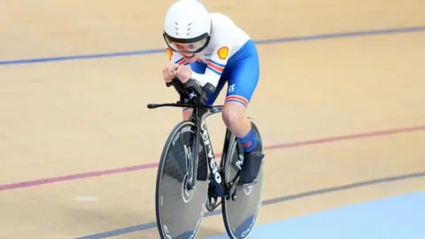 SWPIX Daphne Schrager cycling on a black bike, wearing white and blue, looking straight ahead