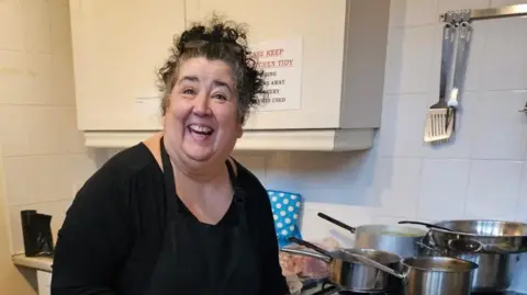 A smiling woman in a black top holding some plates in a kitchen.