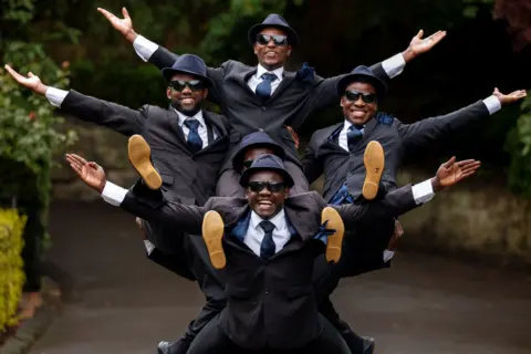 Getty Images The Black Blues Brothers pose during a photocall for their Fringe show. Five men wearing matching dark suits, ties, hats and sunglasses are arranged in a stacked formation, with arms extended outward and legs raised.