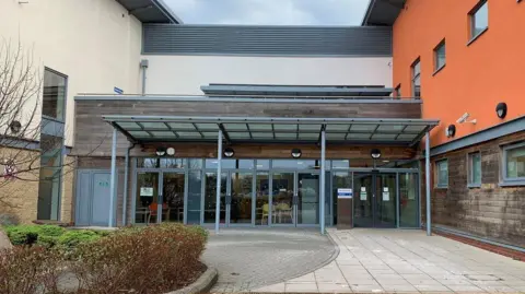 NHS The entrance doors at Frome community hospital, which has orange and wooden-cladding on its outside walls.