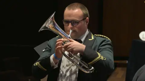 Brass Pass A young man is pictured from the waist up. He is wearing a white shirt and green blazer with gold threaded embellishments. He is holding a silver tenor horn to his mouth.