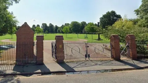 Black gates with brick pillars that open into a park which has a circular walkway and a stretch of green with various trees dotted about.