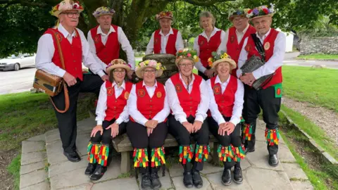 Supplied Eleven members of Furness Morris Dance smiling at the camera back in 2023. They are wearing black trousers, white shirts and red waistcoats, and have hats with flowers at the top.