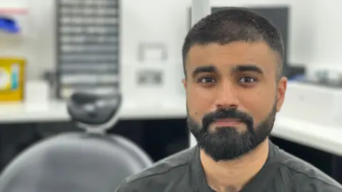 A man with black hair, a beard and brown eyes in a grey shirt sits in a dental surgery. A white counter and black chair can be seen blurred behind him.