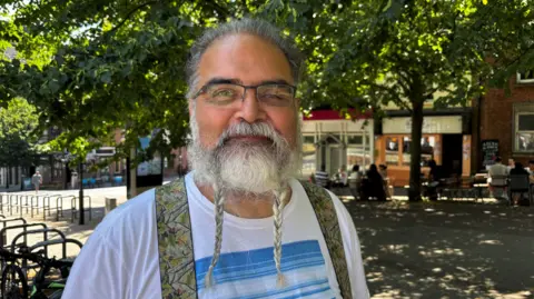 Gemma Dillon Image shows man wearing a white T-shirt with blue stripes stood in the shade of a tree. He has braces on, glasses and his beard is plaited in two braids. 