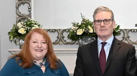 Reuters A man in formal attire with glasses standing beside a woman in a blue top with red hair who is smiling.