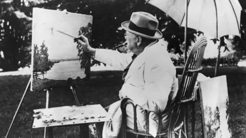 Getty Images A black and white photo of Winston Churchill sitting on a chair, under an umbrella and painting. 