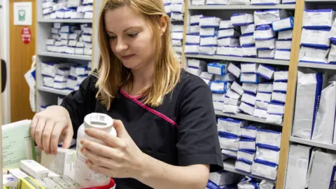 A female pharmacist with blonde hair and a black uniform works in the stock room of a pharmacist