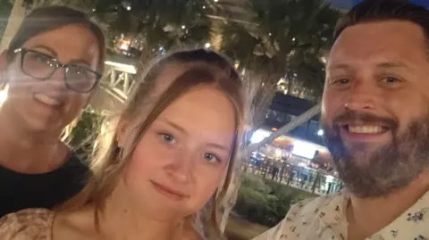 Helen and Lee Draper A young woman is standing in the middle of her mum and dad smiling at the camera. She has a pink dress on and mousey blonde hair. Her dad is on the right and has a white shirt with dark blue small patterns. Her mum is on the left with a black top. It is a head and shoulders selfie shot of them.