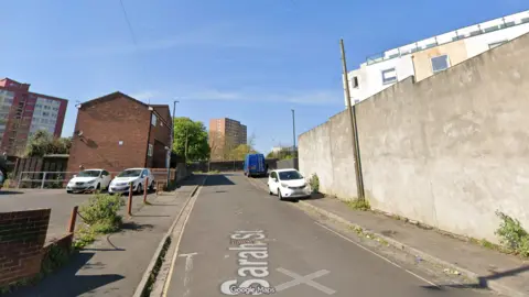 Google Maps A Google street view of Sarah Street in Bristol, it is a short street with a small car park on the left and a tall wall on the right. There are council tower blocks in the background.