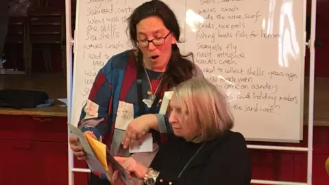 A poet with a care home resident standing in front of a whiteboard with poetry written all over it. It looks like she is reading something out with the resident who is seated.