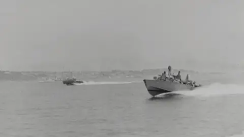 RON 30 boats speeding out of St Helier Harbour - black-and-white photograph showing two motor torpedo boats speeding across open water. Several crew members are visible as small figures on deck. A second boat follows at a distance toward the centre-left, also leaving a wake.