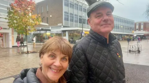 Helen Mulroy/BBC Image shows 66 year old Sadie Mann, with brown hair and wearing a black puffer jacket, smiling at the camera alongside her husband Andy, 68, in a flat cap and padded jacket, they stand in the centre of Bedford's shopping district. 