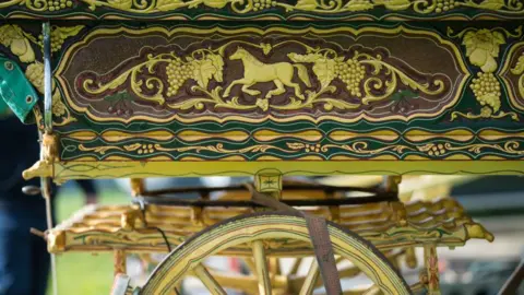 Getty Images A close-up of the wheel and carriage of a traditional travellers' caravan, a horse, grape and leaf motif painted in gold. The caravan appears to be parked in a field, and a man's leg is blurred next to it.