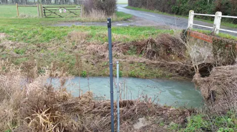 A vertical pole, supporting a tubular sensor, is embedded in the bank of a small stream. Water is running in the background and flowing under a small brick road bridge.