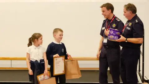 Nottinghamshire Fire and Rescue Service Harry and Grace holding certificates and paper bags. There are two fire service employees wearing navy uniform and holding two boxes of Heroes.