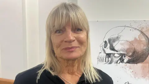 The head and shoulders of an older lady with blonde hair and blue eyes and pink lipstick looking at the camera, she is smiling and wearing a black top and a silver necklace. She is in front of a white background and over her left shoulder there is a picture of a black and white skull