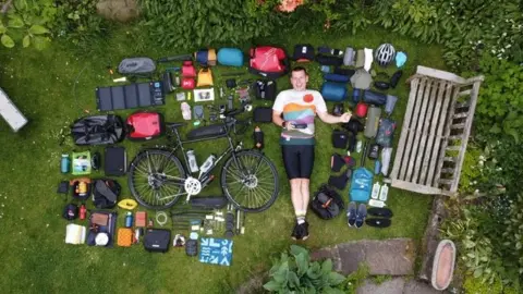 Simon Watts Simon Watts laying down in a garden surrounded by various items laid out next to his bike.