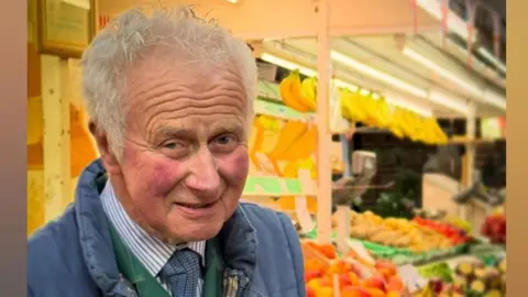 Robin Blair is standing in front of his fruit and vegetable stall and smiling at the camera. He has short white hair and rosy cheeks. He is wearing a blue body warmer and a green jacket underneath.
