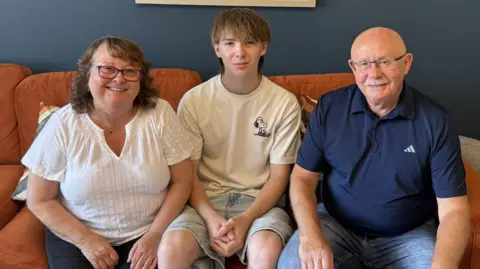 Gateshead Council Jacqui and Malcolm are sitting either side of Jack on a sofa. They are pensioner-age, with Jacqui wearing glasses and having shoulder-length, brown hair. Malcom is also wearing glasses and is bald with a moustache. Jack is a tall teenager with a fringe hairstyle and long hair at the back. The sofa is orange.