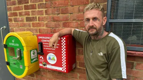 Carl Scott stands beside a bright red emergency bleed control kit mounted on a brick wall, next to a yellow defibrillator box with a green front. The red box has a keypad lock and white text explaining how to access it, including instructions to use an app or call police. Scott wears a short-sleeved green T-shirt and has tattoos on one arm and neck.