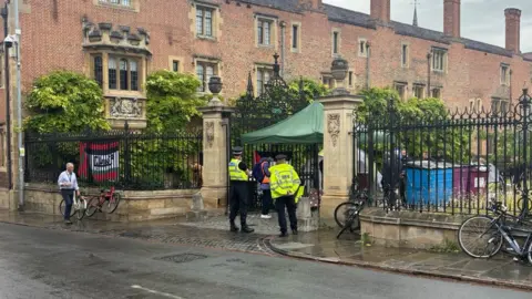 Police outside a college gate talking to masked protesters within and beneath a gazebo