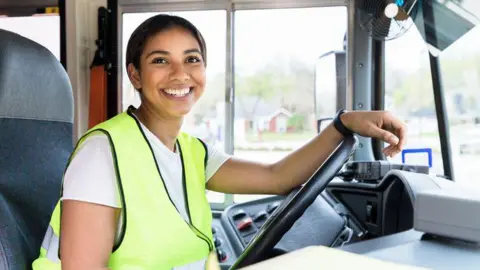 A woman bus driver sits behind the wheel and smiles at the camera. She is wearing a yellow high visibility jacket.