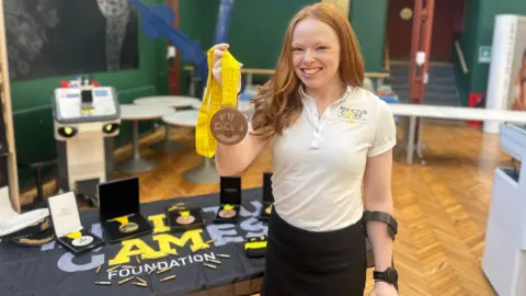 A woman with long ginger hair, wearing a white shirt and black skirt, smiles as she holds a bronze medal. Behind her is a table with other medals on display.