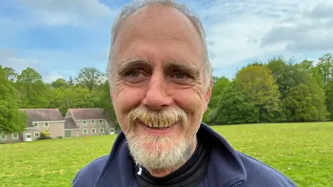 A man with short grey hair and a beard with a blue top in front of a large area of grass