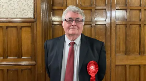 Mike Leach has grey hair and glasses is standing in front of a wood panelled wall. He is wearing a black suit with a white shirt and red tie. He is also wearing a red rosette.