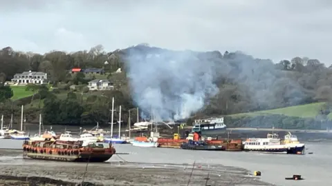 A general view of a creek where boats are moored. It is low tide. Smoke fills the air as the fire continues on the boat. Smoke is fogging up the greenery in the background. Properties are built into the hillside overlooking the water.