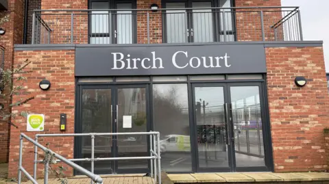 Danny Fullbrook/BBC The main entrance to Birch Court is two sets of glass double-doors. There is an accessible ramp entrance on the left and steps on the right. The name "Birch Court" is displayed on a large grey sign in white font above.