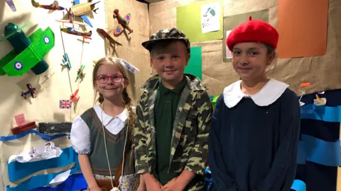 Clare Worden/BBC Evie, Max and Milly dressed in 1940s style clothing in front of a display of D-Day vehicles