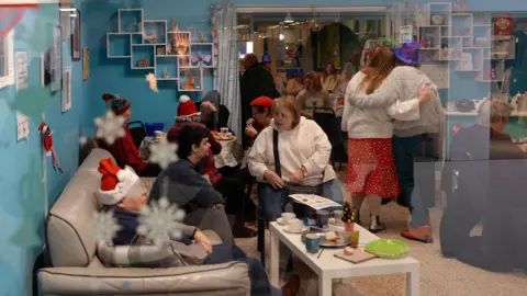 A lively community room decorated for Christmas, with people chatting around sofas and tables, crafts on display, and blue walls lined with photos and shelves.