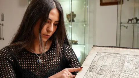 Duggleby Stephenson The photograph shows a dark haired woman wearing a black, long-sleeved, mesh-patterned top, looking down at the open pages of a large, old book. The book has thick, yellowed pages.