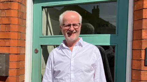 Andrew is standing in front of a green door, smiling at the camera. He is wearing a white shirt and black glasses. He has short white hair and stubble.
