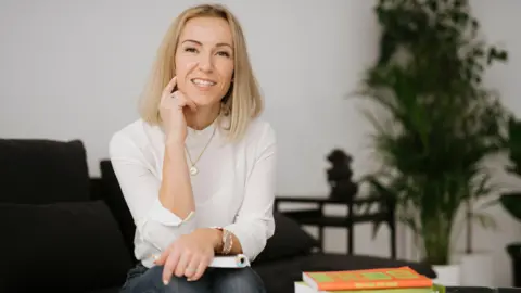 Agata Piatkowska Aleksandra Pamplett sitting with her arms crossed over a book on her lap. She is blonde and wears a white blouse and blue jeans.