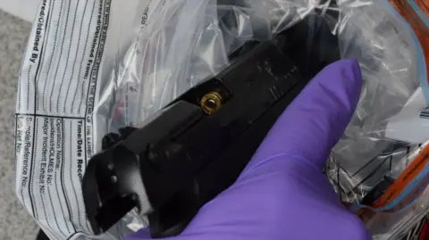 Bedfordshire Police A black handgun held in purple-gloved hand. Its chamber has been opened to reveal the top of a bullet. It is being placed in a plastic evidence bag. 