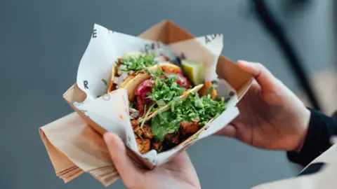 A hand holding tacos in a takeaway container