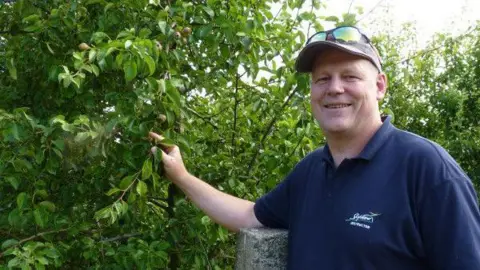 Stephen Ware A man in a blue polo shirt and cap smiles at the camera. He stands next to an apple tree and holds the leaves.