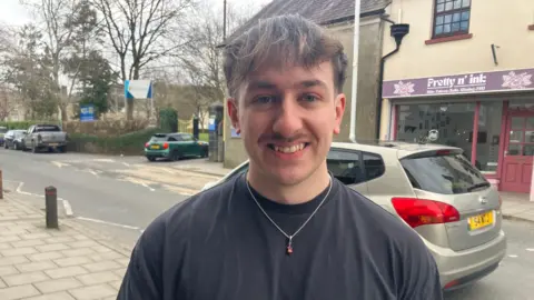 Lewis Jarvis-Blower pictured on the pavement with car on the road behind him and shop window visible on the other side of the road. He wears a black t-shirt and necklace over it and has a moustache and dark short hair.