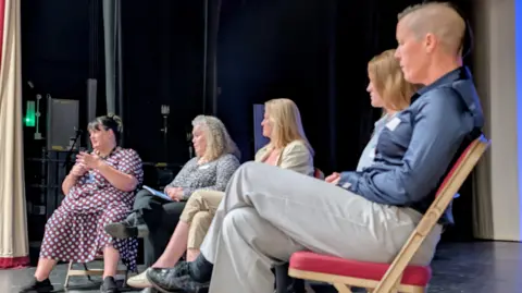 Wiltshire PCC Office Picture of a panel in discussion on a stage, taken from the side. Four people sit on chairs in a semi-circle. One at the end is speaking to the audience.